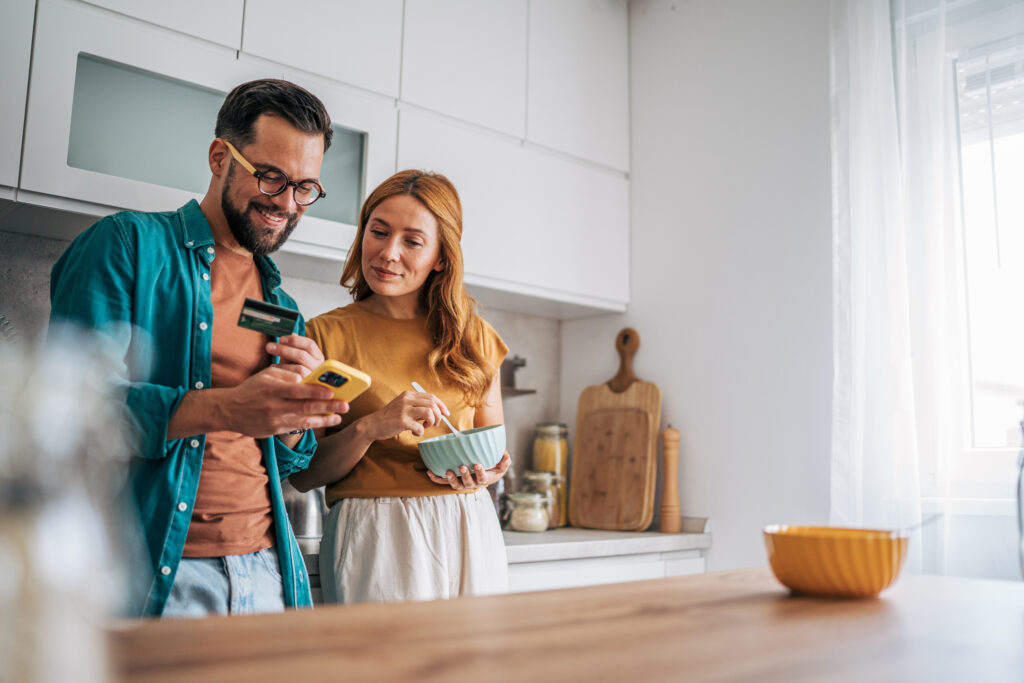Young couple enjoying breakfast in a modern kitchen, using a smartphone and credit card for convenient online grocery shopping, fostering connection and togetherness during their morning routine