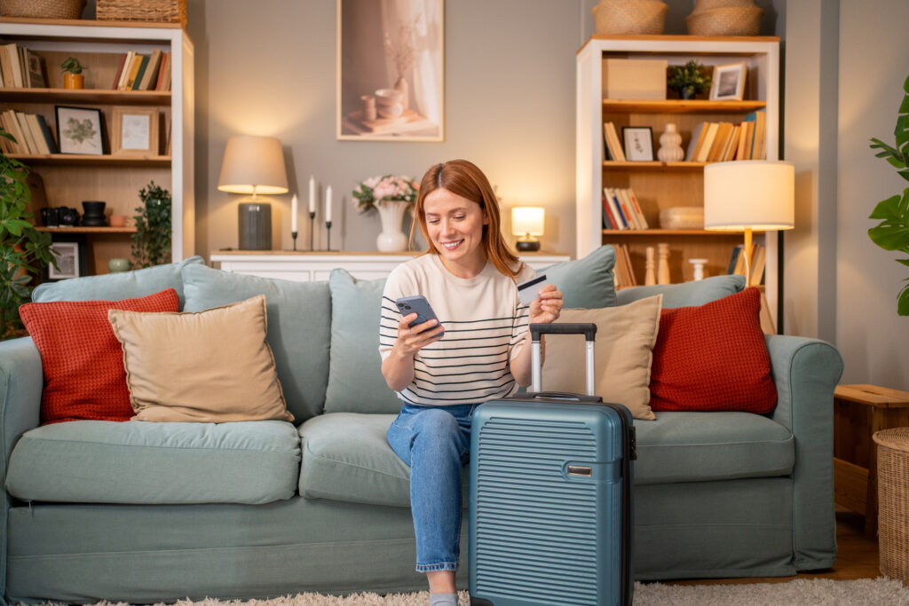 Smiling woman sitting on sofa with suitcase next to her, holding smartphone and credit card, booking travel tickets online, getting ready for vacation.