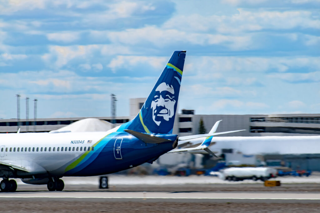 The view of the tail and logo of an Alaska Airlines Boeing 737-800 aircraft landing at the Spokane International Airport under cloudy skies, smoking the tires upon touchdown.
