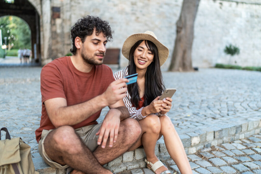 Young couple on a historic cobblestone street completes an online purchase together, man holding a credit card while woman uses a smartphone during their vacation