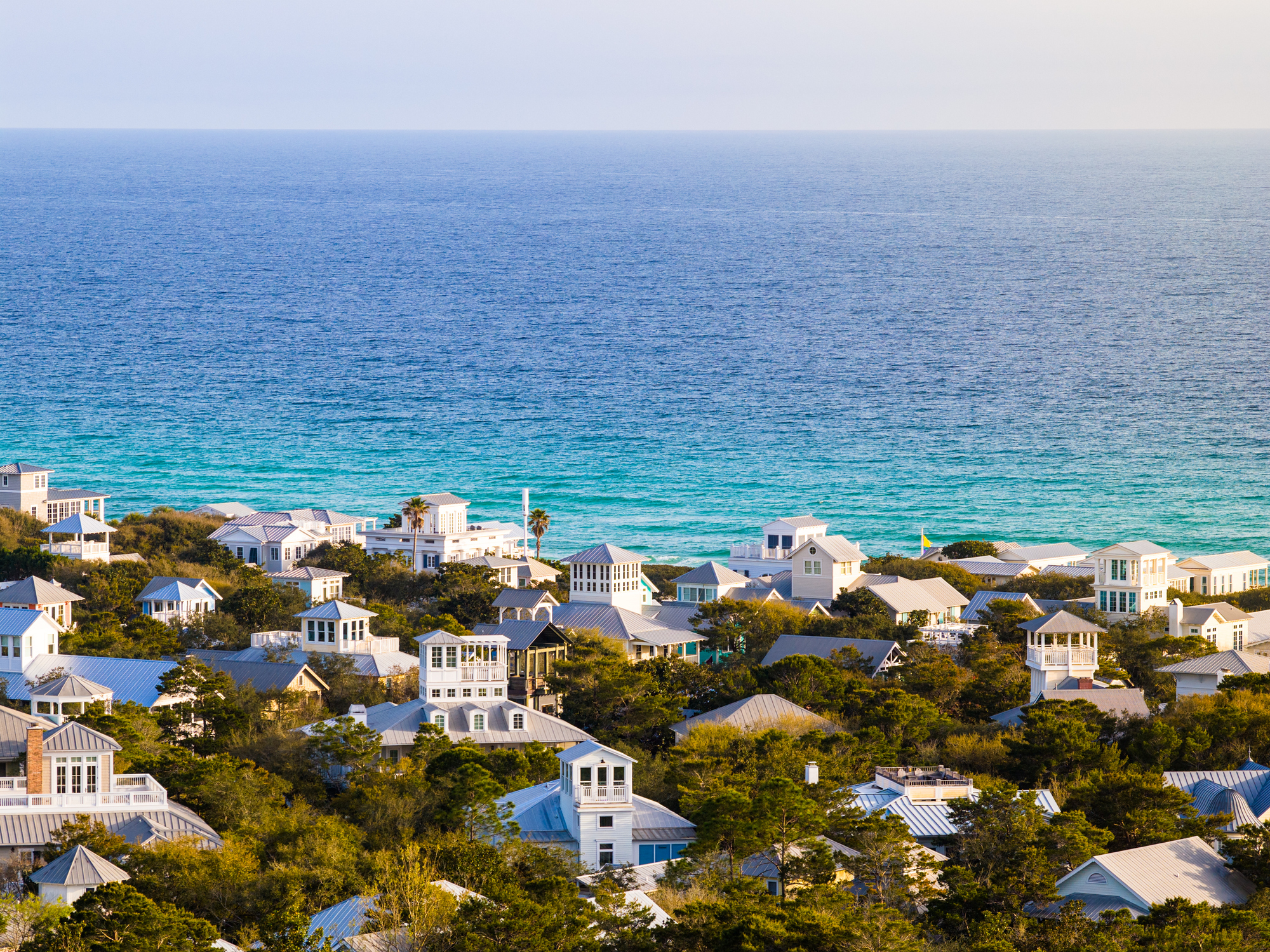 Aerial view of Seaside, Florida beachfront community nestled along the Gulf of Mexico. White and blue residential homes with distinctive architecture are densely arranged among mature trees. Clear turquoise and blue water stretches across the horizon. Spring vegetation is in full growth. Calm water conditions and bright natural lighting suggest midday during pleasant weather.