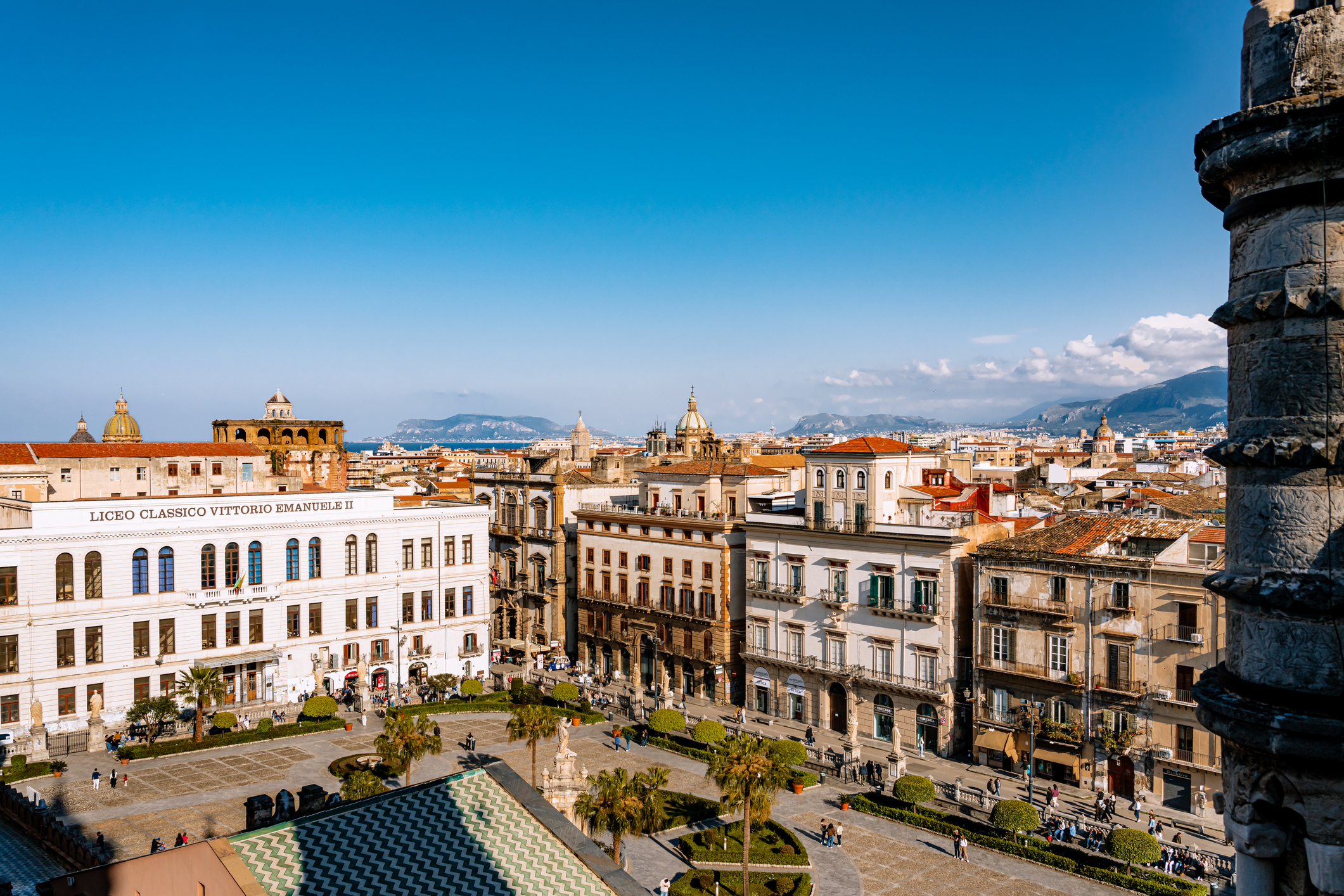 A panoramic view over a busy Palermo city square lined with historic 19th-century buildings, the Liceo Classico Vittorio Emanuele II façade, tiled roofs, distant mountains and clear blue sky on a sunny day.